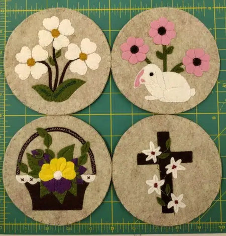 Four round embroidered coasters with floral, bunny, basket, and cross designs on a green cutting mat.